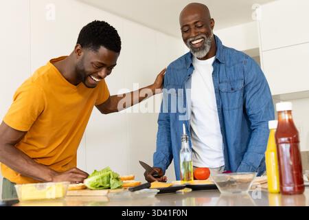 Afroamerikanischer Vater und Sohn, die Burger mit Salat in der Küche zusammenstellen, mit Schneidebrett Stockfoto