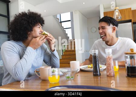 Essen und Lachen, verschiedene männliche Freunde sitzen zu Hause am Esstisch, mit Sandwich und Saft Stockfoto