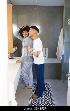Zwei Männer pflegen Haare und umarmen sich am modernen Waschbecken im Badezimmer, mit Spiegelschrank und Badematte Stockfoto