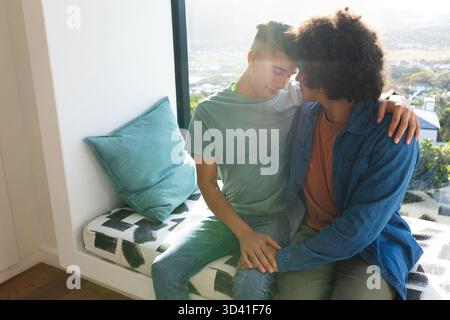 Verschiedene männliche Partner kuscheln sich auf die eingebaute Fensterbank in der Fensterecke, mit blauem Kissen Stockfoto