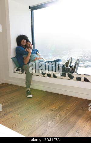 Verschiedene männliche Partner umarmen, die zu Hause auf der Fensterbank sitzen, mit blaublauem Überwurfkissen Stockfoto
