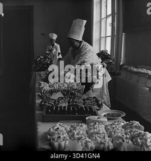 Ein Archivfoto aus dem Jahr 1984 zeigt die Vorbereitungen für ein Buffet im Artyom House of Culture (DK) in Sloviansk, Ukrainische SSR. Dies war für die Delegierten einer Bezirks- und Stadtkonferenz bestimmt. Zwei weibliche Köchinnen in voller Uniform mit hohen Kochmützen arrangieren eine große Aufteilung. Die Tische sind voll mit Eclairs, großen Kuchen und Nelkenvasen. Diese Szene zeigt den „hinter den Kulissen“ eines offiziellen sowjetischen Ereignisses. Dies ist ein Moment des Alltags im friedlichen Donbass vor dem Krieg. Stockfoto