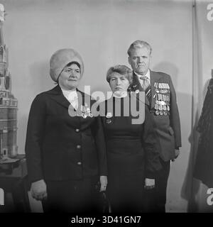 Ein Archivfoto aus dem Jahr 1984 zeigt Delegierte einer Stadtkonferenz im Kulturhaus Artyom in Sloviansk, Ukrainische SSR. Ein Gruppenporträt von drei Teilnehmern: Zwei Frauen und ein männlicher Veteran des Zweiten Weltkriegs. Alle drei sind mit sowjetischen Orden und Orden dekoriert. Die Frau links trägt einen großen Pelzhut, was auf kaltes Wetter hinweist. Sie stehen in der Nähe eines komplizierten Modells, möglicherweise des Kremls. Dies fängt die geehrten Teilnehmer einer offiziellen sowjetischen Veranstaltung ein. Dies zeigt den Alltag im friedlichen Donbass vor dem Krieg. Stockfoto