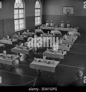 Ein Archivfoto aus dem Jahr 1984 zeigt die Kantine für Delegierte im Artyom House of Culture (DK) in Sloviansk, Ukrainische SSR. Diese hohe Aussicht zeigt das Fitnessstudio mit einem Basketballkorb und markiertem Boden, das in einen Speisesaal umgewandelt wurde. Die Tische sind mit weißen Tischdecken, Mahlzeiten und Milchflaschen aus Glas besetzt. Vier Köche stehen in Uniform an der Buffetlinie. Das ist das Catering für eine offizielle sowjetische Veranstaltung. Dies ist ein Moment des Alltags im friedlichen Donbass vor dem Krieg. Stockfoto