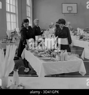 Ein Archivfoto aus dem Jahr 1984 zeigt Kellnerinnen, die sich auf ein Bankett im Artyom House of Culture (DK) in Sloviansk, ukrainische SSR, vorbereiten. Die Konferenzkantine wurde in der Turnhalle eingerichtet, wie der Basketballkorb zeigt. Die Kellner in dunklen Kleidern und weißen Schürzen haben Tische mit Tellern, Obst und Glasmilchflaschen gedeckt. Im Hintergrund stehen Köche in Kochmützen an der Buffetzeile. Das ist das Catering für eine offizielle sowjetische Veranstaltung. Dies ist ein Moment des Alltags im friedlichen Donbass vor dem Krieg. Stockfoto