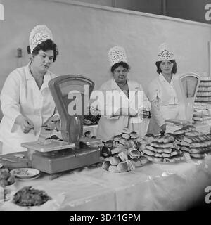 Ein Archivfoto aus dem Jahr 1984 zeigt das Buffet-Personal des Artyom House of Culture (DK) in Sloviansk, ukrainische SSR. Drei Arbeiterinnen in weißen Mänteln und dekorativen Spitzenhüten posieren während einer Konferenz hinter einem Tisch. Der Tisch ist mit frischen Brötchen, Gebäck und anderen Snacks bedeckt. Zwei Sätze mechanischer Waagen sind bereit, um Teile für die Teilnehmer zu wiegen. Das ist das Catering für eine offizielle sowjetische Veranstaltung. Dies ist ein Moment des Alltags im friedlichen Donbass vor dem Krieg. Stockfoto