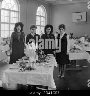 Ein Archivfoto aus dem Jahr 1984 zeigt vier Delegierte auf einer Bezirks- und Stadtkonferenz in Sloviansk, ukrainische SSR. Die Frauen posieren für ein Porträt hinter ihrem Tisch während einer Mittagspause. Die Kantine wurde zeitweise im Turnsaal des Kulturhauses Artyom (DK) eingerichtet, was an den Bogenfenstern und dem Basketballkorb zu erkennen ist. Der Tisch ist mit Tellern mit Speisen, Obst und Getränken gedeckt. Dies fängt die Atmosphäre eines offiziellen sowjetischen Ereignisses ein. Dies ist ein Moment des Alltags im friedlichen Donbass vor dem Krieg. Stockfoto