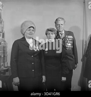 Ein Archivfoto aus dem Jahr 1984 zeigt Delegierte einer Stadtkonferenz im Kulturhaus Artyom in Sloviansk, Ukrainische SSR. Ein Gruppenporträt von drei Teilnehmern: Zwei Frauen und ein männlicher Veteran des Zweiten Weltkriegs. Alle drei sind mit sowjetischen Orden und Orden dekoriert. Die Frau links trägt einen großen Pelzhut, was auf kaltes Wetter hinweist. Sie stehen in der Nähe eines komplizierten Modells, möglicherweise des Kremls. Dies fängt die geehrten Teilnehmer einer offiziellen sowjetischen Veranstaltung ein. Dies zeigt den Alltag im friedlichen Donbass vor dem Krieg. Stockfoto