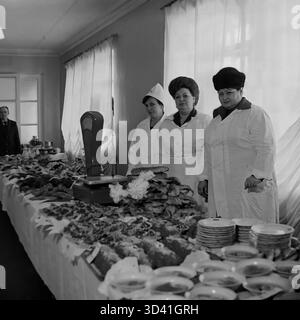 Ein Archivfoto aus dem Jahr 1984 zeigt die Buffetlinie für eine Konferenz im Artyom House of Culture (DK) in Sloviansk, Ukrainische SSR. Drei weibliche Küchenangestellte in weißen Uniformen und Hüten posieren hinter einem langen Tisch. Der Tisch ist voll mit einer großen Auswahl an kalten Vorspeisen („zakuski“), Fleisch, Salaten und Brot, mit Waagen und Stapeln sauberer Teller, die für die Teilnehmer bereit sind. Das ist das Catering für eine offizielle sowjetische Veranstaltung. Dies ist ein Moment des Alltags im friedlichen Donbass vor dem Krieg. Stockfoto