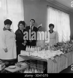 Ein Archivfoto aus dem Jahr 1984 zeigt einen männlichen Delegierten (im Anzug, mit Abzeichen) und weibliche Mitarbeiter bei einer Konferenz im Artyom House of Culture (DK) in Sloviansk, Ukrainische SSR. Sie posieren hinter einem Buffettisch. Der Tisch ist voll mit Backwaren, Brötchen und zahlreichen Glasmilchflaschen. Drei Frauen sind in Küchenuniform. Das ist das Catering für eine offizielle sowjetische Veranstaltung. Dies ist ein Moment des Alltags im friedlichen Donbass vor dem Krieg. Stockfoto