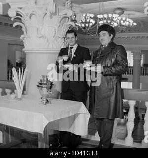 Ein Archivfoto aus dem Jahr 1984 zeigt zwei männliche Delegierte bei einer Konferenz im Kulturhaus Artyom (DK) in Sloviansk, Ukrainische SSR. Die Männer sind auf einer Teepause, stehen an einem Tisch mit einem traditionellen Samovar. Sie befinden sich im großen Foyer, erkennbar an seinen verzierten Säulen und Kronleuchtern. Der Mann auf der rechten Seite trägt einen markanten Pelzhut (Uschanka), was darauf hindeutet, dass es kalt ist. Dies fängt einen Moment der Diskussion während eines offiziellen sowjetischen Ereignisses ein. Dies zeigt den Alltag im friedlichen Donbass vor dem Krieg. Stockfoto