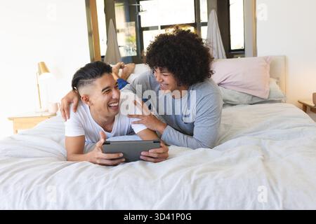Lachende männliche Freunde, die auf dem Bett in einem modernen Schlafzimmer liegen und ein Tablet neben Lampe und Kissen halten Stockfoto