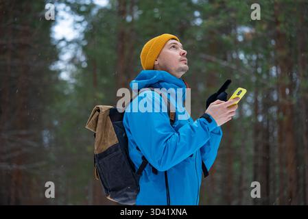 Junger Mann in Winterkleidung und Rucksack, der sich in einem schneebedeckten Wald verirrt, ein Smartphone für die Navigation benutzt und nach Orientierung in der tiefen Natur Ausschau hält Stockfoto