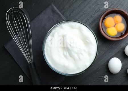 Eiweiß in Schüssel, Schneebesen und Zutaten auf schwarzem Tisch, flach gelegt Stockfoto