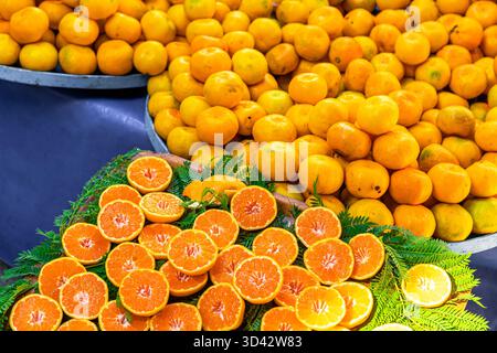 Frische Orangen und Mandarinen auf einem Markt. Ganze Orangen sind hoch gestapelt, während geschnittene Orangen auf einem Bett aus grünen Blättern angeordnet sind. Stockfoto