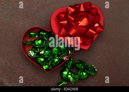 Rote Geschenkbox in Herzform mit glänzender Schleife und grünen Bonbons auf dunkler strukturierter Oberfläche mit Kopierraum. Elegantes und festliches Stillleben für Chr Stockfoto