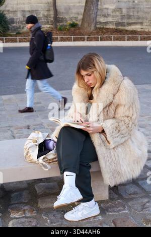 Junge Frau, die im Winter ein Buch in einem gemütlichen Pelzmantel auf einer Pariser Straße liest, Paris, Frankreich Stockfoto