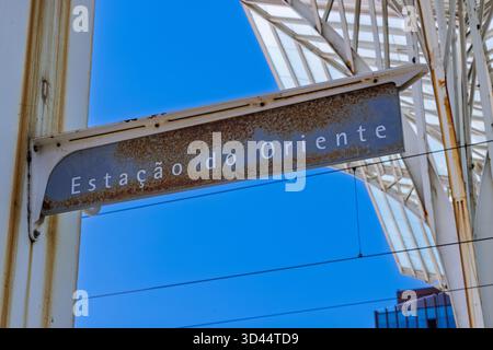 Nahaufnahme des Schilds „Estação do Oriente“ auf einer rostigen Metallplatte, eingerahmt vom hellblauen Himmel und der markanten modernen Dachkonstruktion des Bahnhofs. Stockfoto