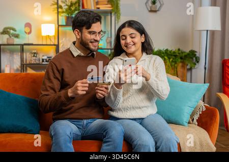 Glückliches indisches Paar beim Online-Shopping, indem es die Kreditkartennummer auf dem Smartphone eingibt, während es zu Hause auf dem Sofa sitzt. Lächelnde Familie ist mit einer guten Aktion für Waren in der Wohnung zufrieden. Stockfoto