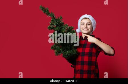 Fröhlicher Junge mit Weihnachtsmann-Hut und rotem kariertem Hemd, der die Daumen hoch hält, ein Zeichen der Anerkennung wie ein kleiner künstlicher Weihnachtsbaum und darauf auf rotem Ba zeigt Stockfoto