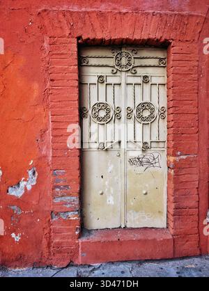 Verzierte Vintage-Metalltür mit kreisförmigem Eisen in einer verwitterten roten Stuckwand mit Zeichen von abblätternder Farbe und Graffiti Stockfoto
