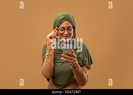 Positive muslimische Frau mit Kopftuch nutzt Smartphone, um neue App vor beigem Hintergrund zu erkunden Stockfoto