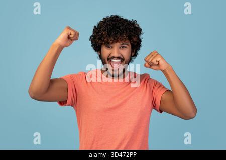 Junger bärtiger Indianer feiert Leistung mit erhobenen Fäusten auf blauem Hintergrund Stockfoto