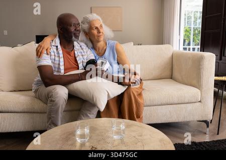Diverse Seniorenpaare, die den Blutdruck im Wohnzimmer überprüfen, mit digitalem Blutdruckmessgerät Stockfoto