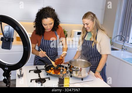 Kochen Sie verschiedene weibliche Freunde, rühren Sie Gemüse in der Pfanne auf der Kücheninsel, mit Ringlicht Stockfoto