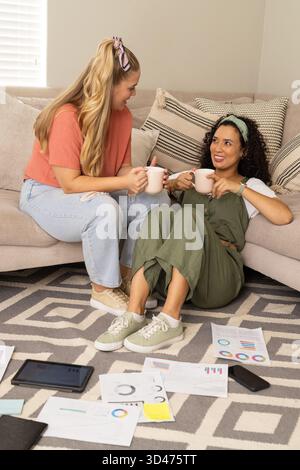 Sitzende Freundinnen, die Karten untersuchen und aus Keramikbechern im Wohnzimmer trinken, mit Tablet Stockfoto
