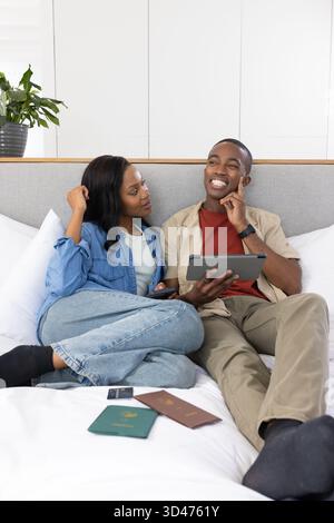 Afroamerikanisches Paar, das sich im Schlafzimmer auf das Bett gestützt hat, mit Tablet, Pässen und Smartphones Stockfoto