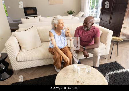 Medikamenteneinnahme diverse Seniorenpaare sitzen auf Sofa im Wohnzimmer, mit Pillen-Flaschen und Wasser Stockfoto