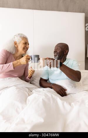 Verschiedene Seniorenpaare sitzen auf dem Bett im Schlafzimmer und halten graue und beige Keramikbecher Stockfoto