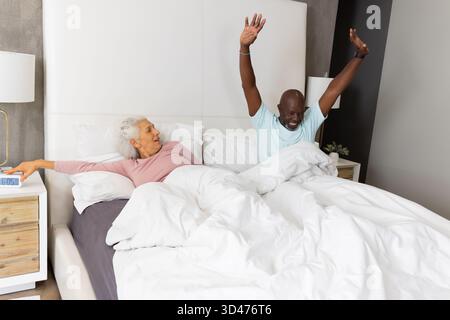 Das reißende Seniorenpaar erhebt sich unter weißer Bettdecke im Bett und greift nach einem Wecker Stockfoto