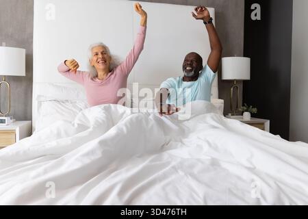 Verschiedene ältere Ehepaare, die sich die Arme ausstrecken, während sie im Schlafzimmer auf einem Kingsize-Bett sitzen, mit Tischlampen Stockfoto