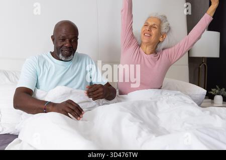 Verschiedene Seniorenpaare, die zu Hause im Bett sitzen, Arme dehnen, mit Bettdecke und Lampe Stockfoto