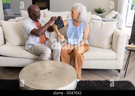 Diverse Seniorenpaare, die den Blutdruck auf dem Sofa im Wohnzimmer überprüfen, mit digitalem Monitor Stockfoto