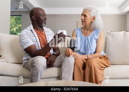 Blutdruckkontrolle bei älteren Ehepaaren mit digitalem Monitor mit Manschette auf beigefarbenem Sofa im Wohnzimmer Stockfoto
