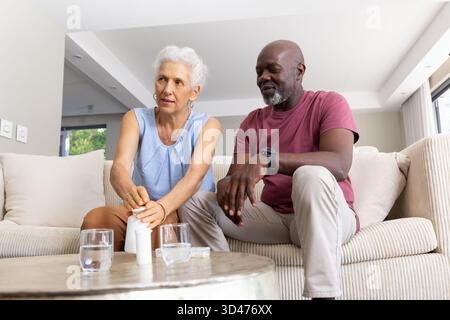 Aufklappbare Pille Flasche diverse Seniorenpaare sitzen auf cremefarbenen Sofa im Wohnzimmer, mit Wassergläsern Stockfoto