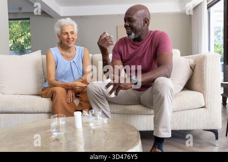 In der Hand der Pillen-Flasche, diverse Seniorenpaare diskutieren Medikamente im Wohnzimmer, mit Wassergläsern Stockfoto