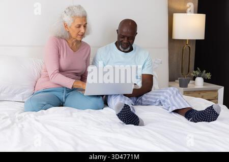 Verschiedene Seniorenpaare, die auf dem silbernen Laptop im Schlafzimmer sitzen, mit Nachttischlampe Stockfoto