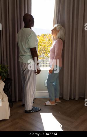 Verschiedene Seniorenpaare, die zu Hause durch graue Vorhänge blicken, mit Blick auf den Balkon und Topfpflanzen Stockfoto