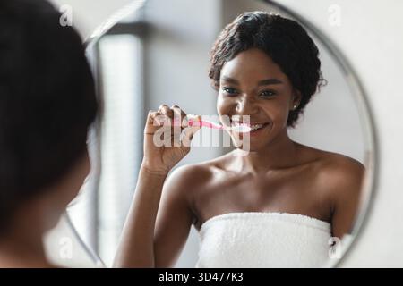 Die junge Schwarze Frau genießt morgens Routine, während sie ihre Zähne vor dem Spiegel putzt Stockfoto