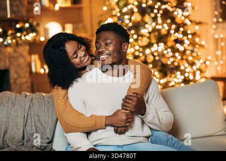 Liebevolles junges schwarzes Paar, das fröhliche Momente an Weihnachten in einem gemütlichen, dekorierten Wohnzimmer teilt Stockfoto
