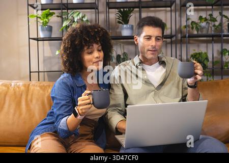 Das Paar sitzt eng zusammen und benutzt einen Laptop auf einem Ledersofa, hält graue Tassen Stockfoto