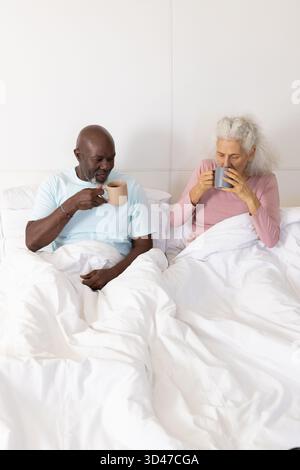 Verschiedene Paare sitzen aufrecht im Schlafzimmer und trinken heiße Getränke aus Keramikbechern Stockfoto
