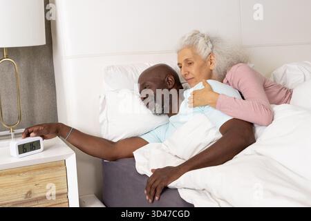 Das Seniorenpaar umarmt und schaltet den digitalen Wecker auf dem hölzernen Nachttisch im Schlafzimmer aus Stockfoto