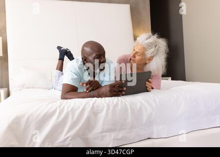Ein älteres Paar, das sich auf einem strahlend weißen Bett mit Tablet und Nachttischlampe zurücklehnen kann Stockfoto