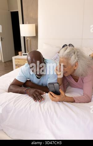 Entspannendes vielseitiges Seniorenpaar mit Blick auf das Smartphone im Schlafzimmer auf dem Bett, mit Lampe und Kaffeetasse Stockfoto
