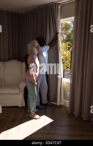 Verschiedene ältere Ehepaare öffnen hellgraue Vorhänge im Wohnzimmer, mit weißem Polstersessel Stockfoto